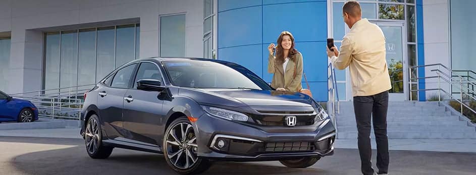 Man in a beige jacket photographing a smiling woman beside a parked black Honda sedan.