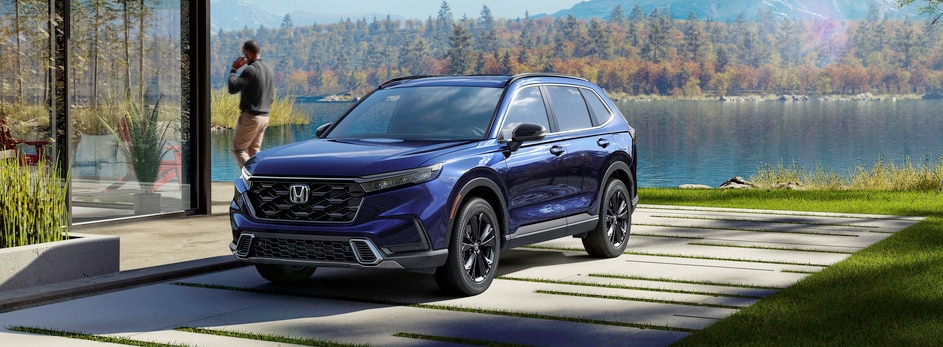 A blue Honda SUV parked outside a modern house, near a lake with mountains and autumn trees in the background.