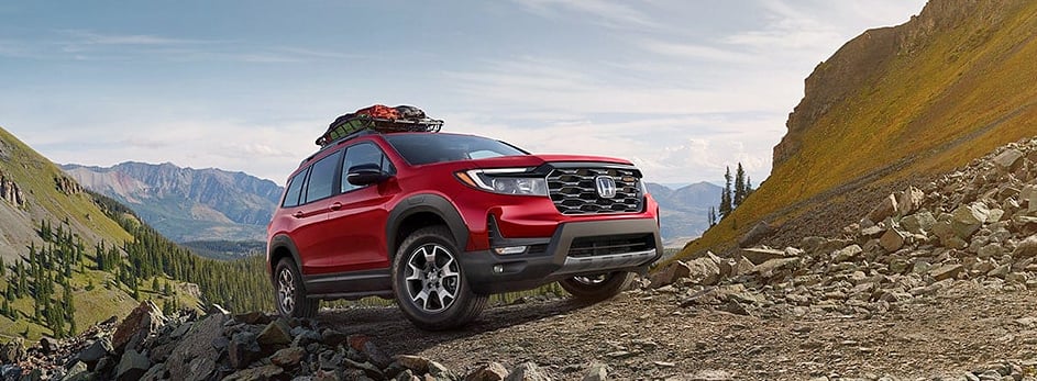 Red SUV on a rocky mountain path with gear on the roof, surrounded by scenic mountainous landscape.