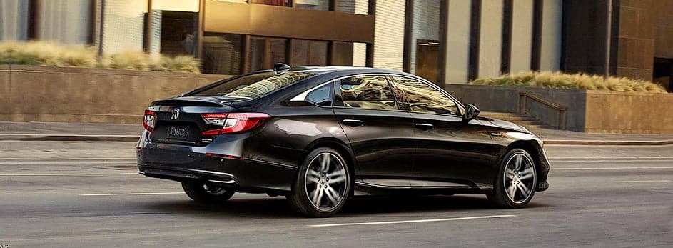 Black sedan car driving on a city street with modern buildings in the background.