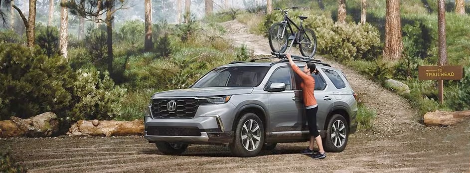 SUV with a bike on the roof rack in a forest setting near a trailhead sign.