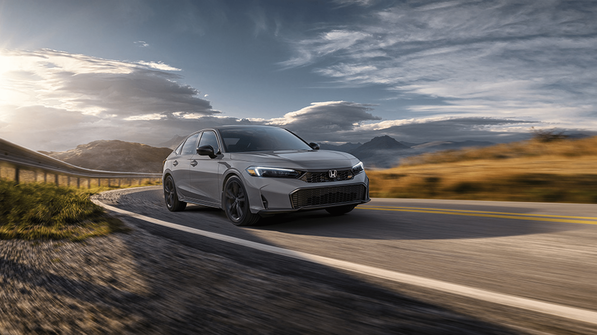 A gray Honda car driving on a scenic mountain road with dramatic clouds and the sun setting in the background.