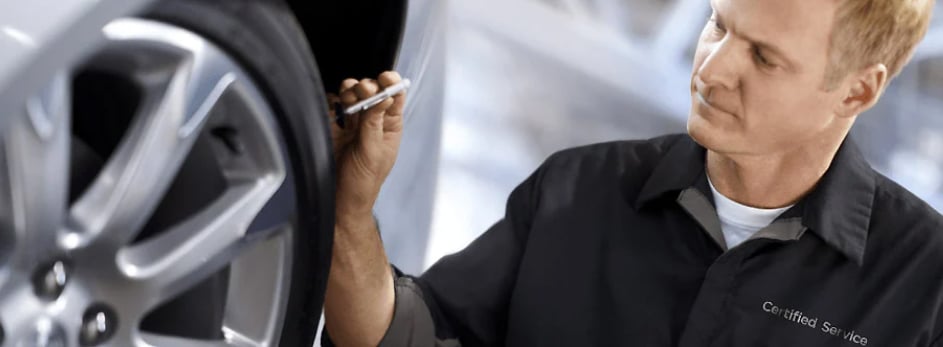 Mechanic in uniform inspecting a car tire, focusing with a pen in hand, labeled "Certified Service."
