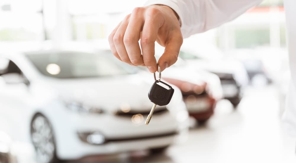 Hand holding car key in dealership, blurred cars in background, symbolizing new car purchase.