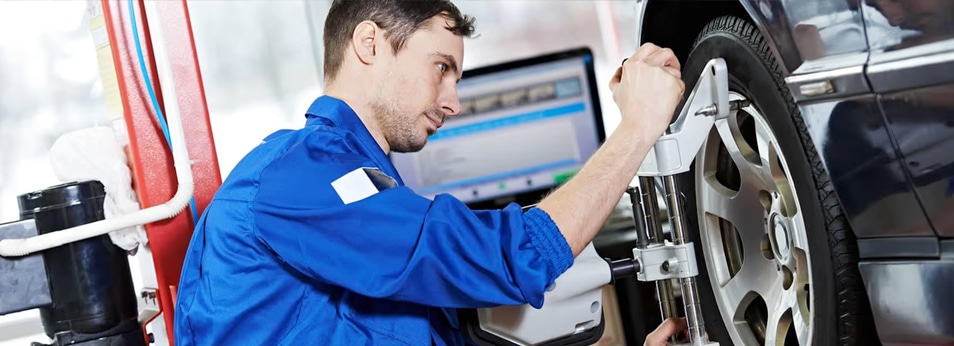 Mechanic in blue uniform adjusting car wheel alignment in a garage with computer equipment.