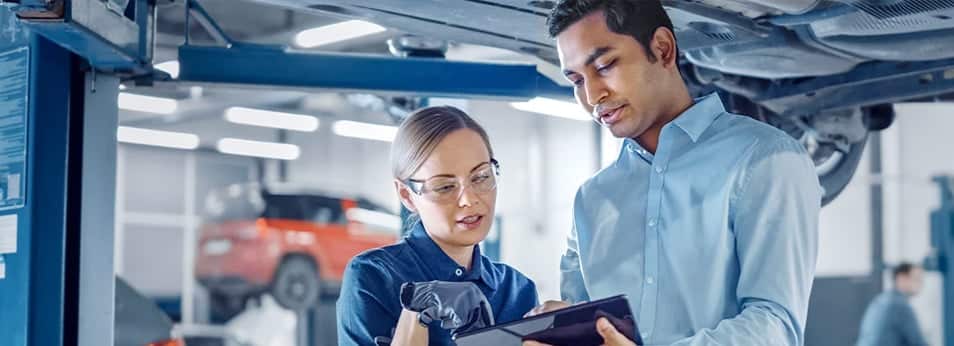 Two mechanics in a car workshop discuss diagnostics with a tablet under a vehicle on a lift.